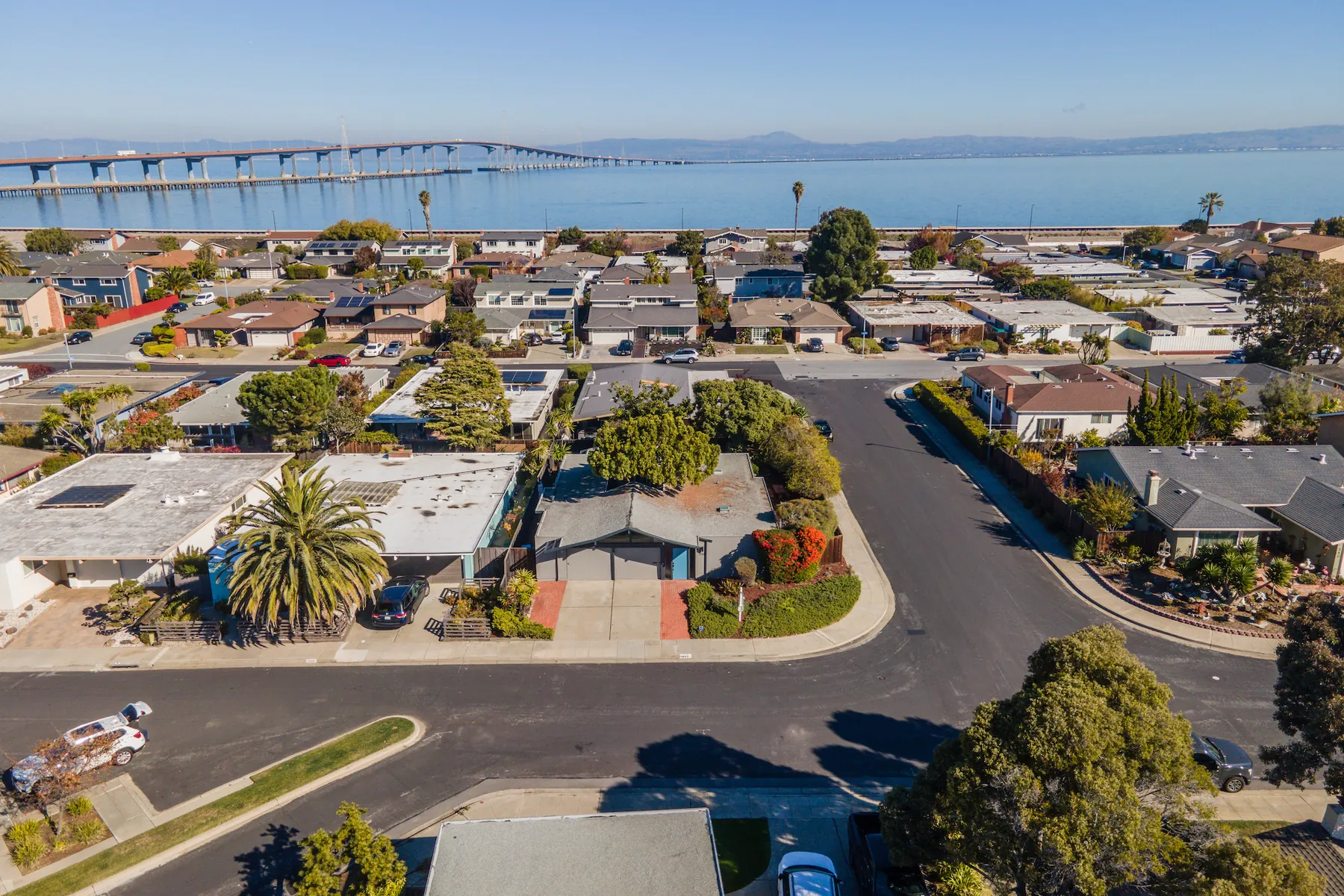 Drone photograph over Foster City rooftops looking north-east across San Francisco Bay, with the San Mateo-Hayward Bridge crossing the upper part of the frame and the levee running along the shoreline behind the last row of houses.