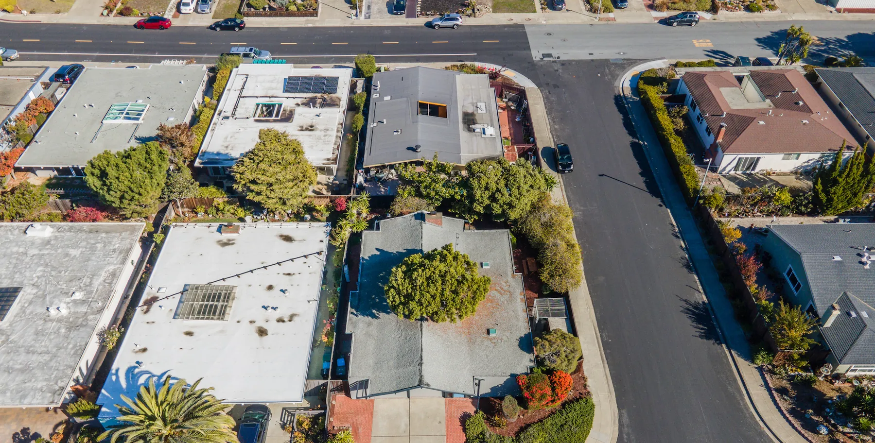 Drone photograph looking straight down on a Foster City block. Two broad flat white roofs and a low grey gabled roof sit side by side in the foreground; across the street the houses have steeper pitched and tiled roofs of a different building type.