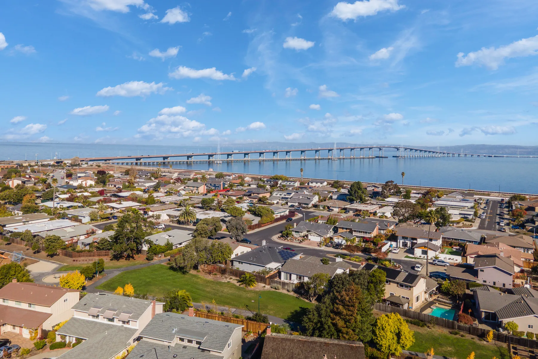 High drone photograph over Foster City looking across San Francisco Bay with the San Mateo-Hayward Bridge spanning the water and rooftops of mixed flat and pitched roofs below.