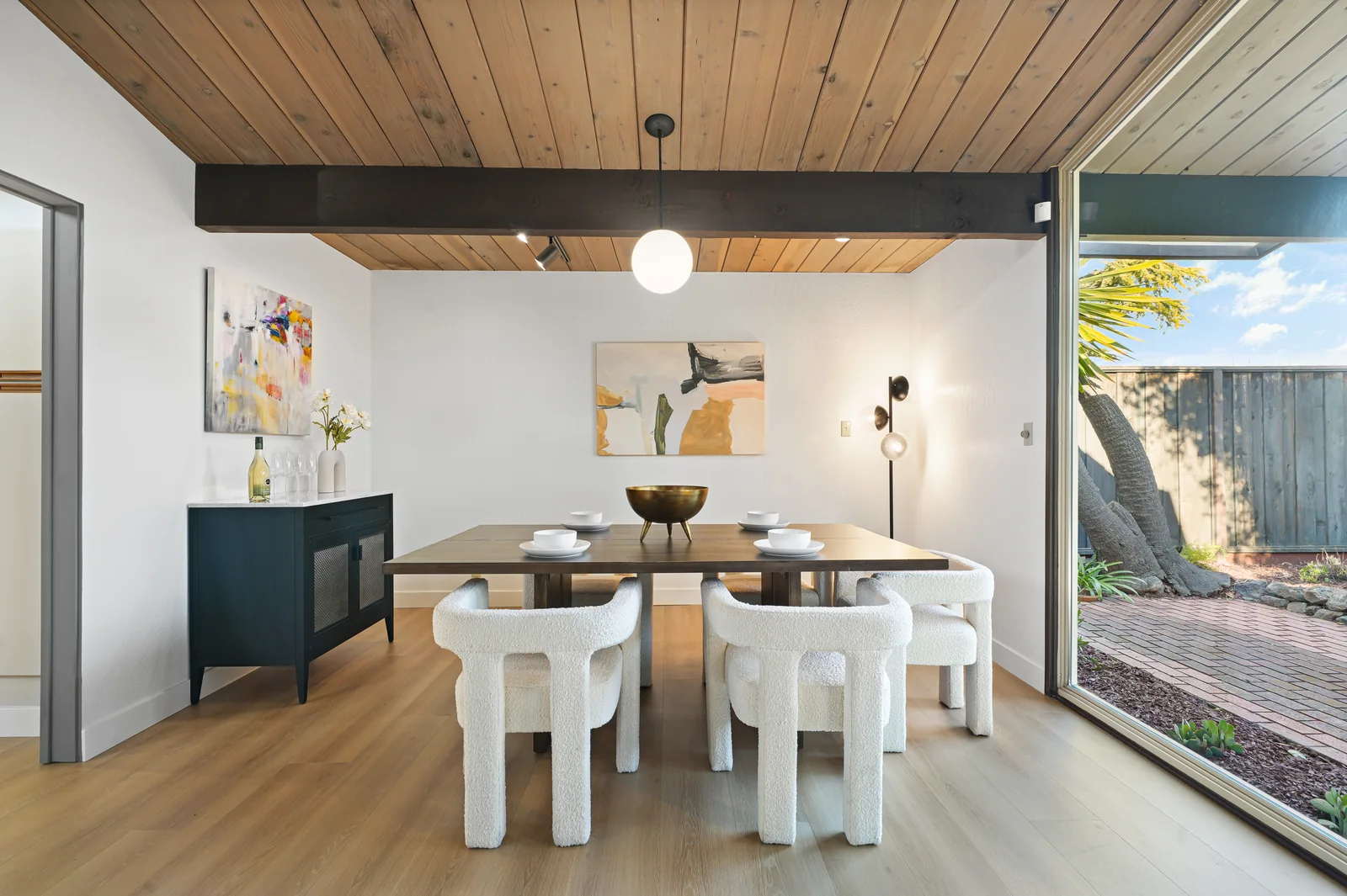 Dining room with the same beamed wood ceiling, a single dark beam crossing it, and a full-height sliding glass wall opening to a brick side yard with a leaning palm.