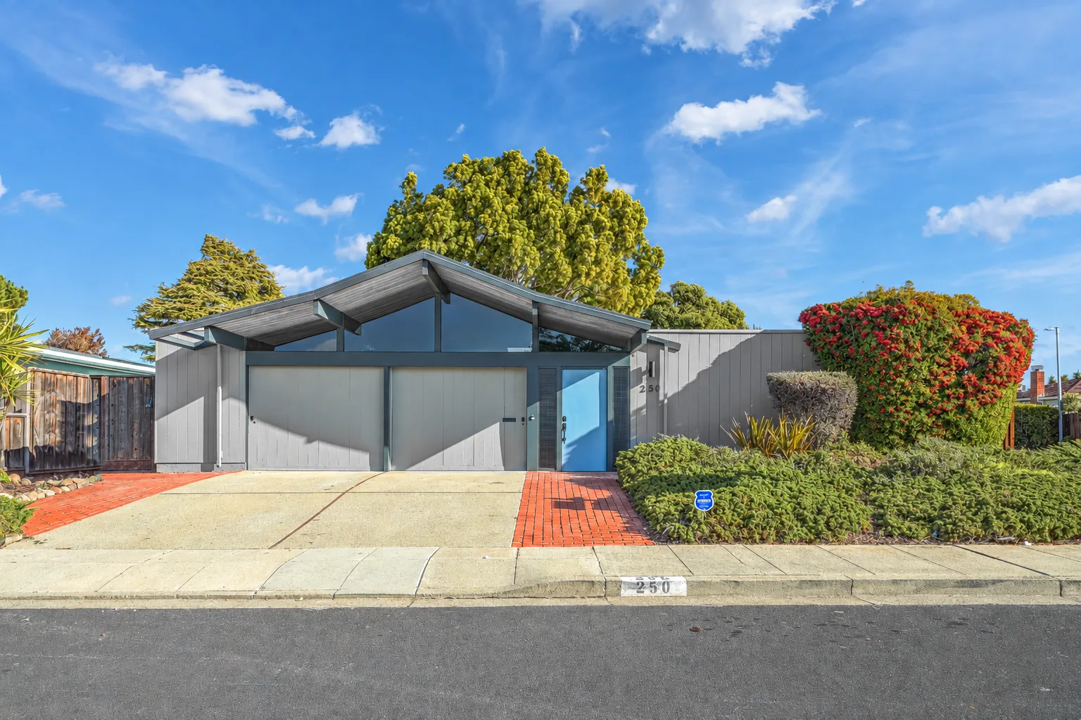 Street elevation of a Foster City Eichler: a low double gable over a deep grey fascia, two grey garage doors, a pale blue front door set behind a perforated metal screen, brick entry path and clipped junipers, with a flowering bottlebrush at the kerb.