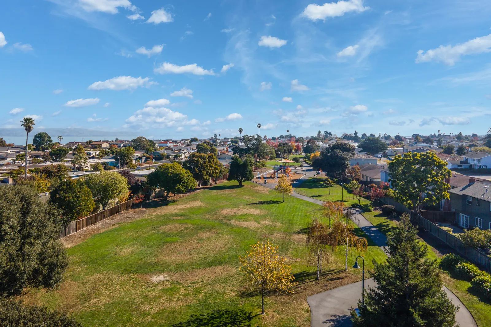 Drone view over a greenbelt lawn and path with houses either side and San Francisco Bay visible on the horizon.