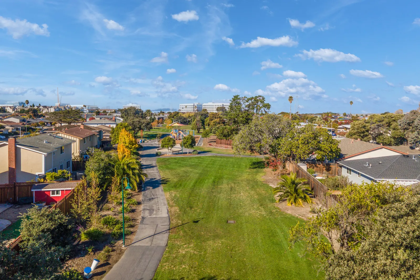Drone view along a landscaped greenbelt with a path, lawn and palms, houses either side and office buildings on the horizon.