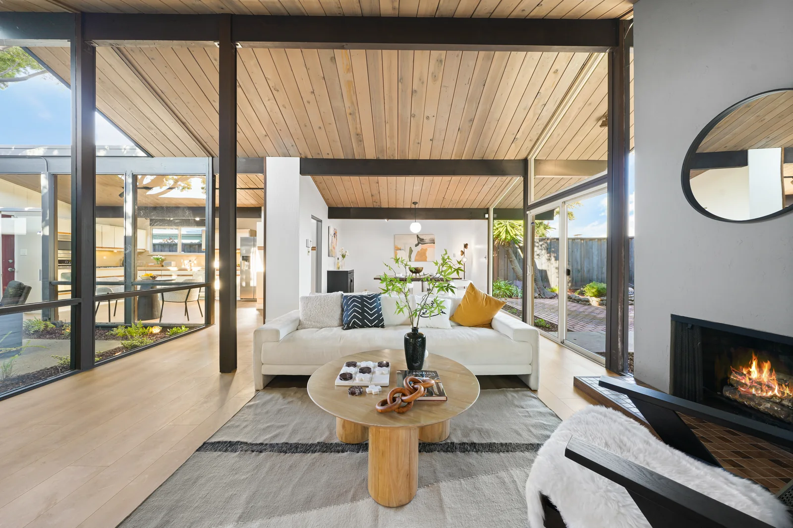 Living room of a Foster City Eichler: tongue-and-groove wood decking laid across exposed dark beams forms the ceiling, glazing runs full height on two sides to a brick terrace, and a fireplace stands against a plastered chimney breast.