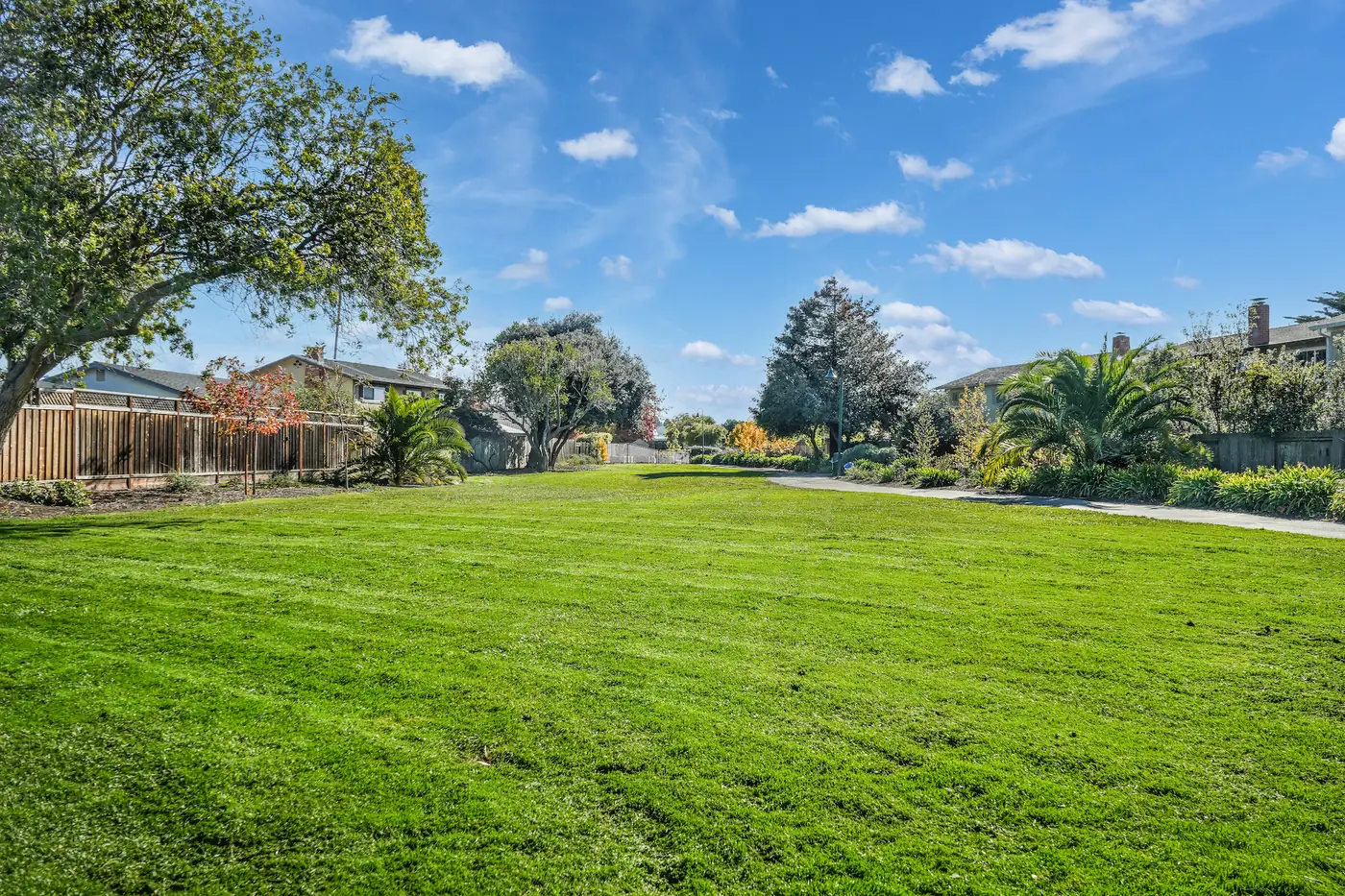 A wide open lawn bordered by wooden fences and mature trees, with two-storey houses beyond.