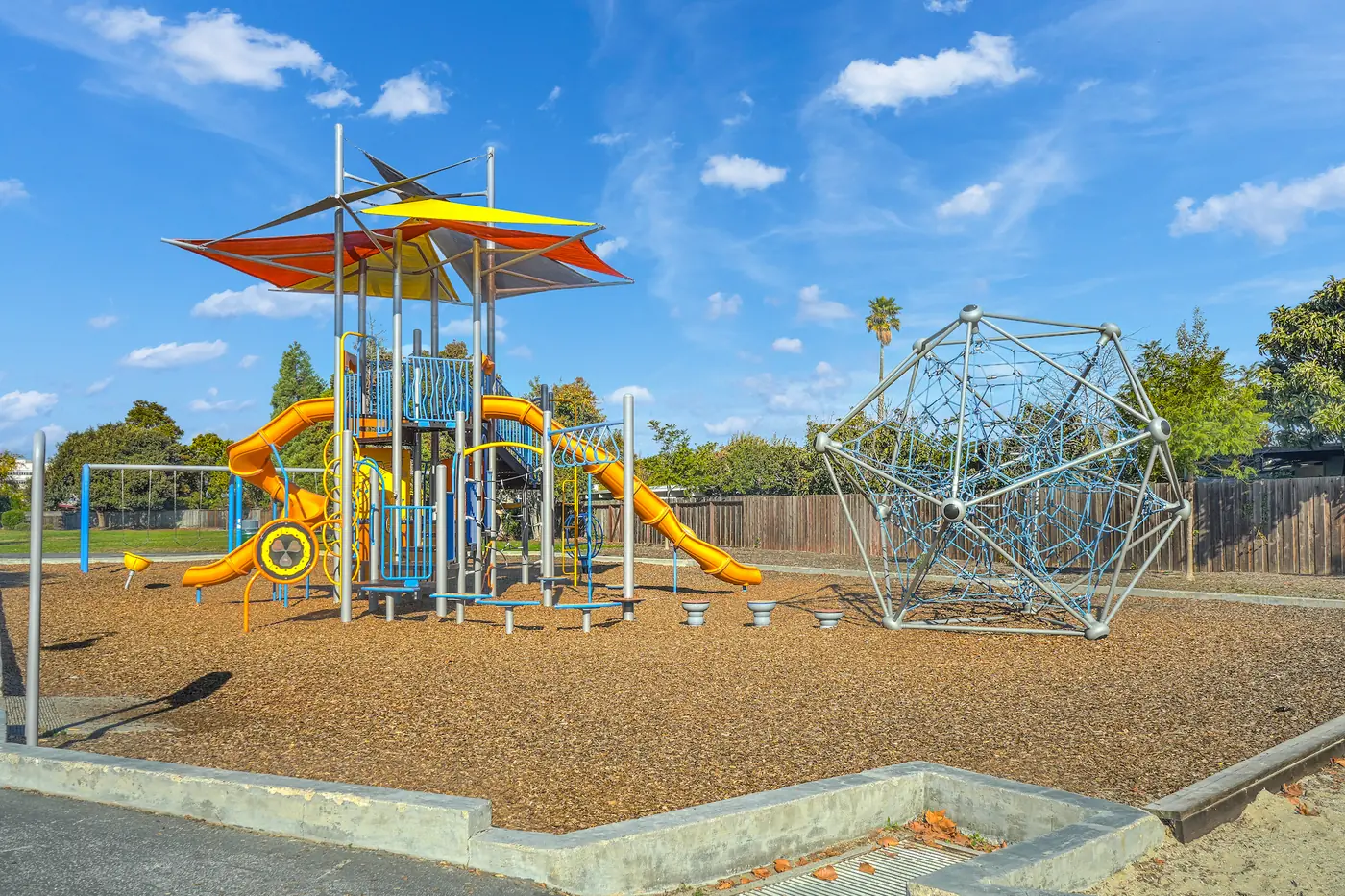 A children's playground with orange slides, a climbing frame and shade sails over bark chippings.